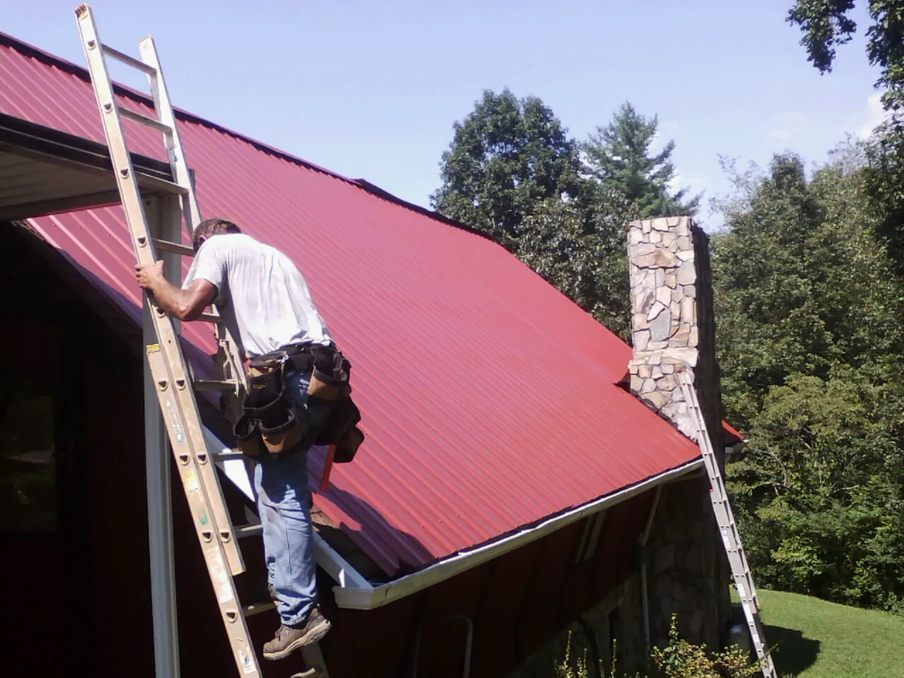 Roofer on a ladder beside a red metal roof and stone chimney — Soffit & Fascia Repair by Roofing Brothers in Ponchatoula